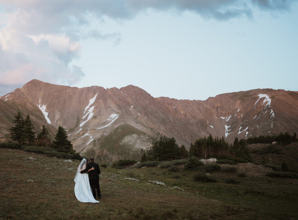 The couple is walking towards large mountain views. they have their arms wrapped around each other in the green grass. The mountains are pink from the sunset. They are leaning into each other. They are outside of Breckenridge at a mountain pass.