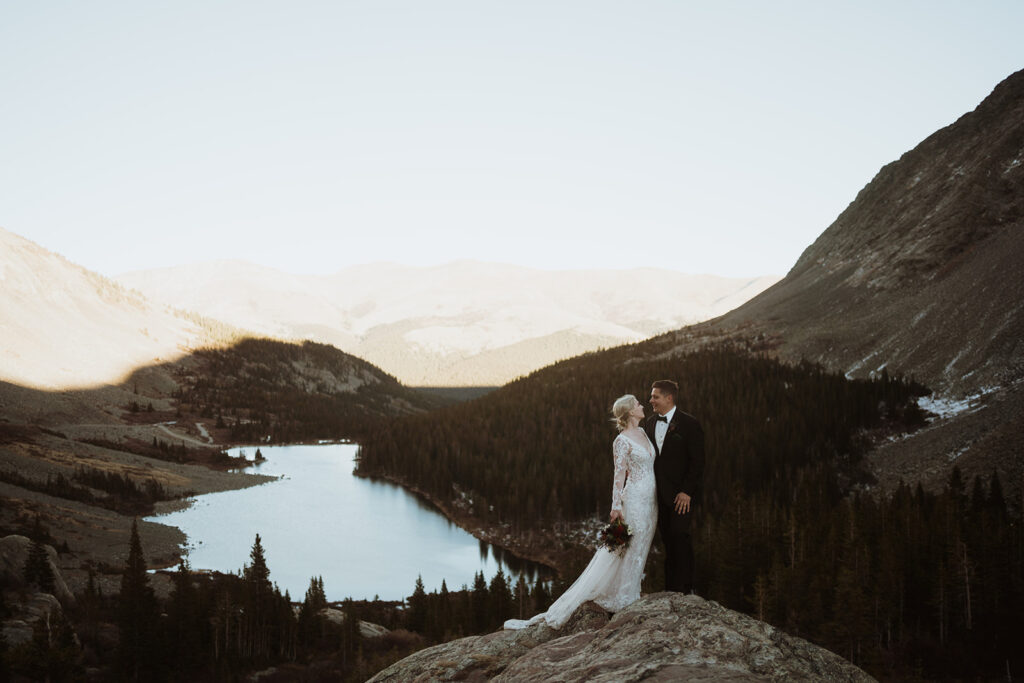 couple is holding onto each other on the edge of a cliff. the bride is holding a bouquet of flowers by her side. the groom is smiling at the bride. there is a lake in the background and mountains that are washed out by the sun.