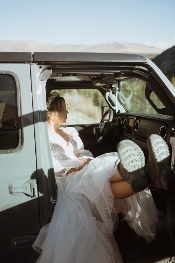 bride is sitting in a jeep and looking out the front of it. Her feet are up against the arm rest and she has a straight face. she is in a white wedding dress and the jeep is a blueish gray. There are mountains in the background.