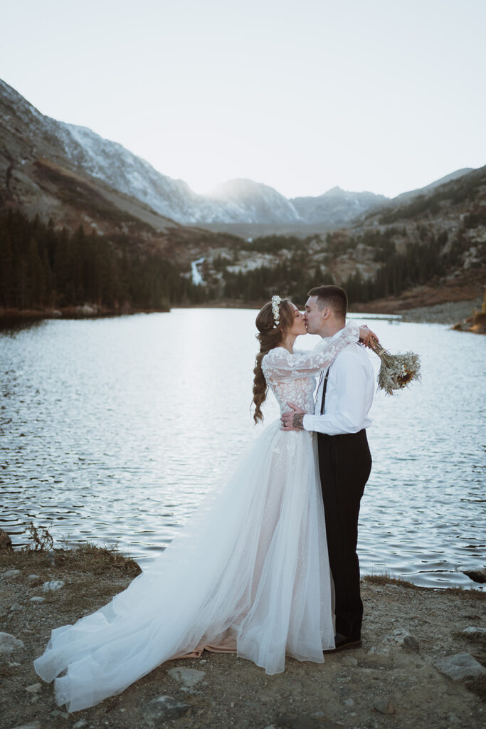 bride and groom are kissing. the bride has her arms wrapped around the groom's neck with flowers in her hands. there is a lake and mountains with waterfalls in the background.