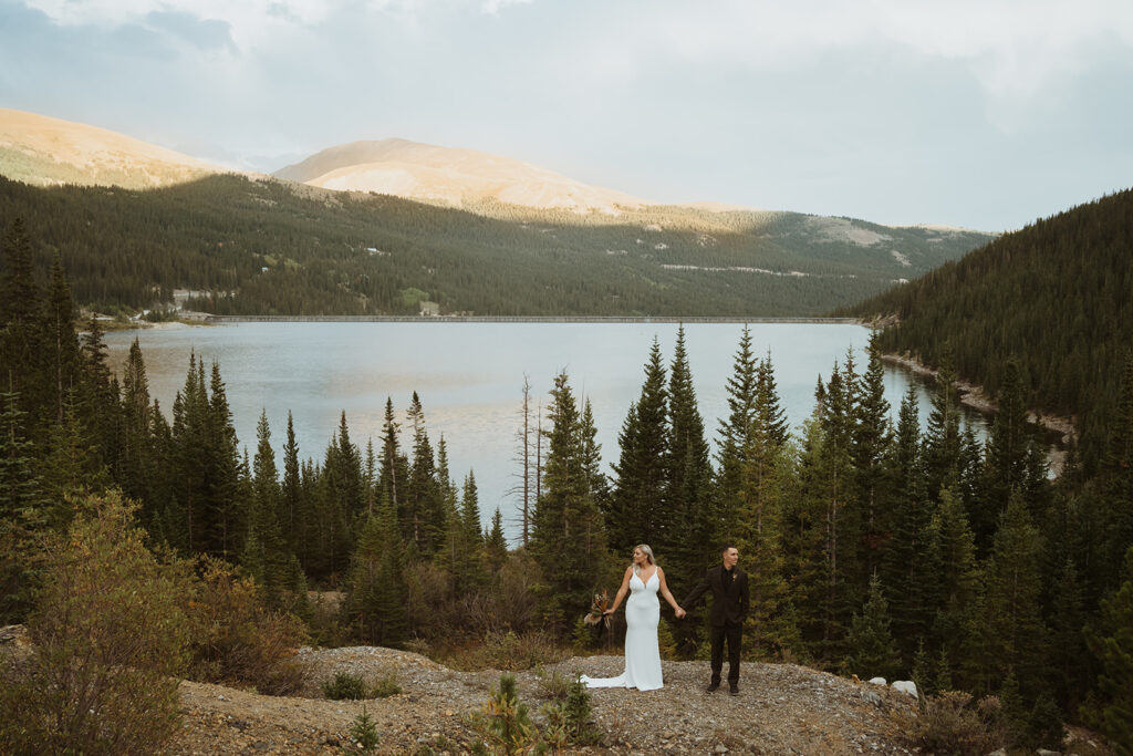Couple is standing in the mountains holding hands about a step apart on a ledge. The bride is in a white dress and the groom is in a black suit. The sun is behind them, and there is a large lake and mountains in the background. There are pine trees and a shoreline.