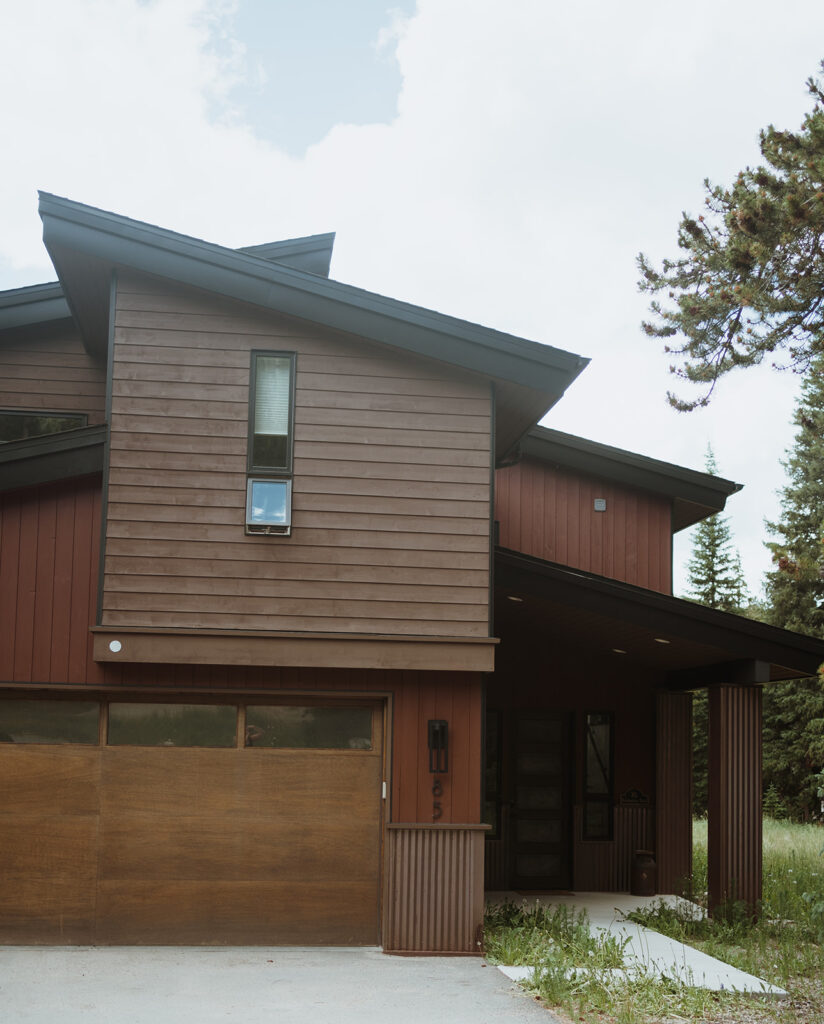 Image of a cabin in Breckenridge. It is painted brown with a brown garage door and red trim. It is a modern looking cabin with a driveway and trees behind it.