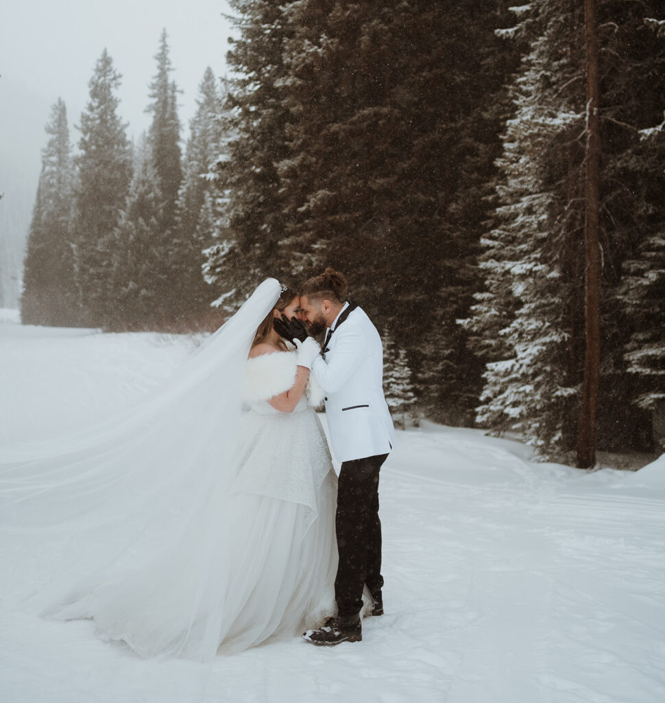 The bride and groom are standing in the snow in winter park for their elopement day. The groom has black gloves on, and is holding the bride's face. The bride has a veil flowing and has on white gloves. She is holding the groom's arms.
