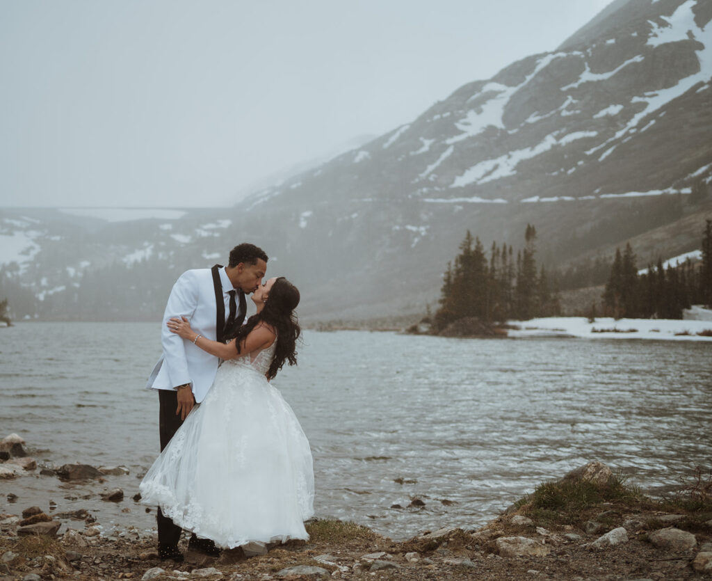 bride is holding onto the groom as they kiss with an alpine lake in the background. there is snow falling and fog surrounding the mountains. They are having a winter elopement in colorado and are going to sign their marriage license at the lake.