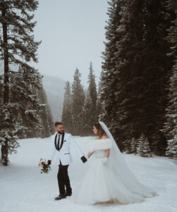 The groom is leading the bride up the cliff edge. She is holding her wedding dress and there are snowy mountains and snow under their feet. 