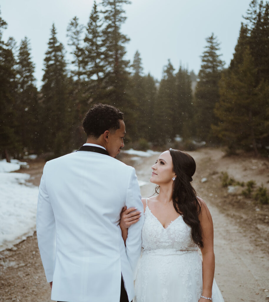 The groom is wearing a white suit with black trim. The bride is in a white wedding dress. The groom is facing backwards towards the trail, and the bride has her arm wrapped around his facing the camera. They are looking at each other as snow falls all around them.