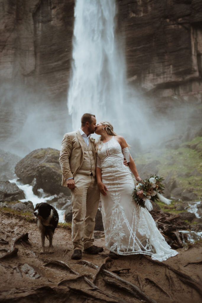The bride and groom are kissing beneath a waterfall. The groom has on a tan suit, and the bride is wearing a lacey white wedding dress. She is holding flowers and they are standing shoulder to shoulder.