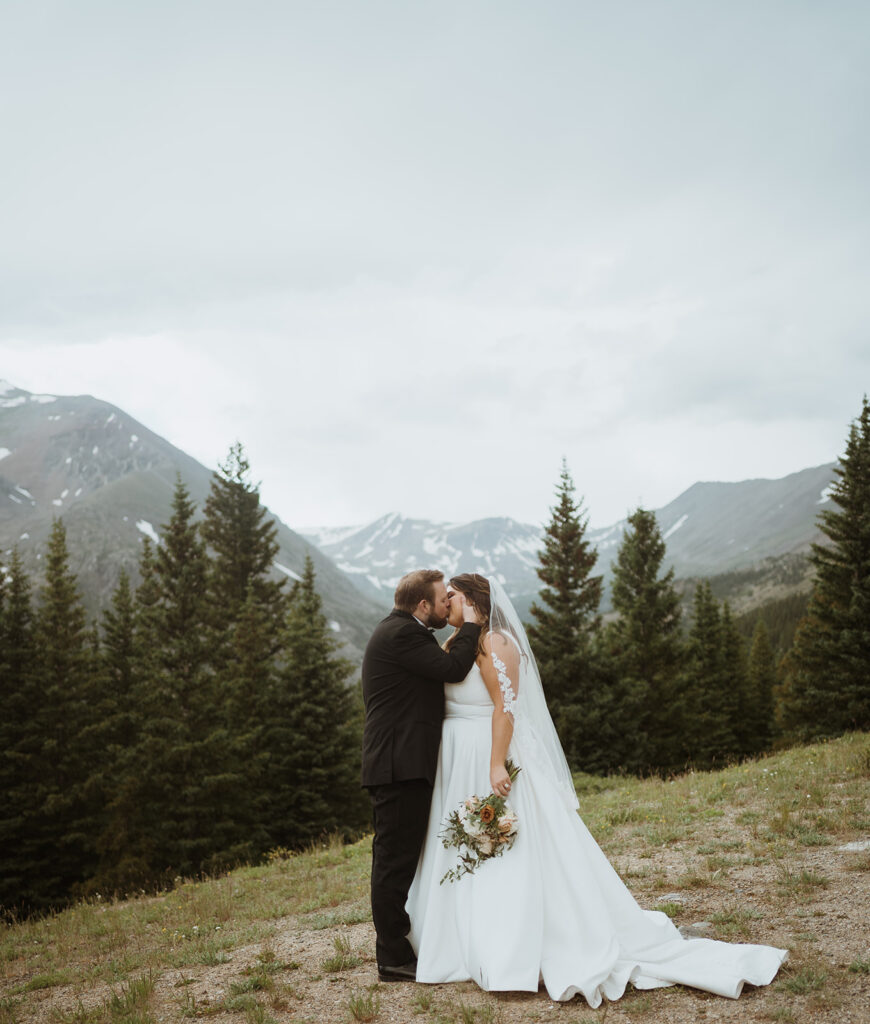 The bride and groom are kissing with mountains in the background. They are having a winter elopement in Colorado, and the bride has her arm down by her side holding her flowers. There are green trees and green grass.