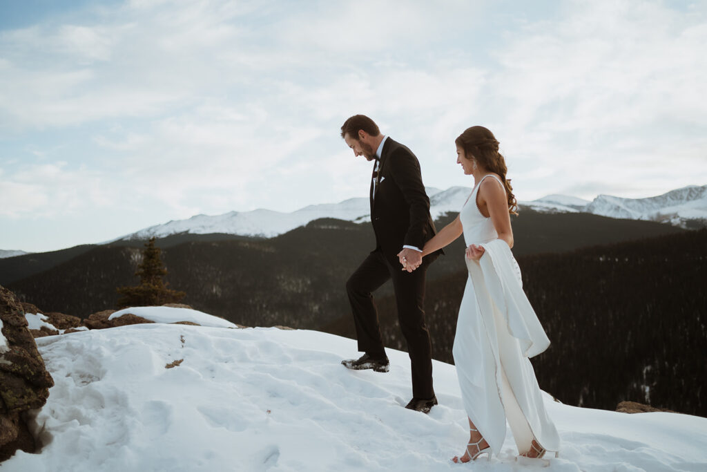 The groom is leading the bride up the cliff edge. She is holding her wedding dress and there are snowy mountains and snow under their feet. 
