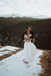 couple is standing in their wedding attire in the snow. The groom is in a black suit and the bride is in a white dress. They are on a cliff edge with mountains in the background and snow on the ground. The groom has the bride wrapped up in his arms.