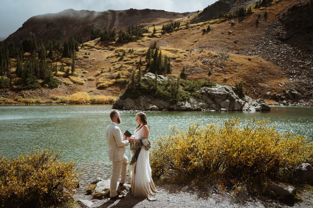 Couple is standing at the base of an alpine lake reading their vows. the bride is holding her vow book and the groom is holding her hands. he is in a tan suit and she is in a white wedding dress with brown on the bottom. 