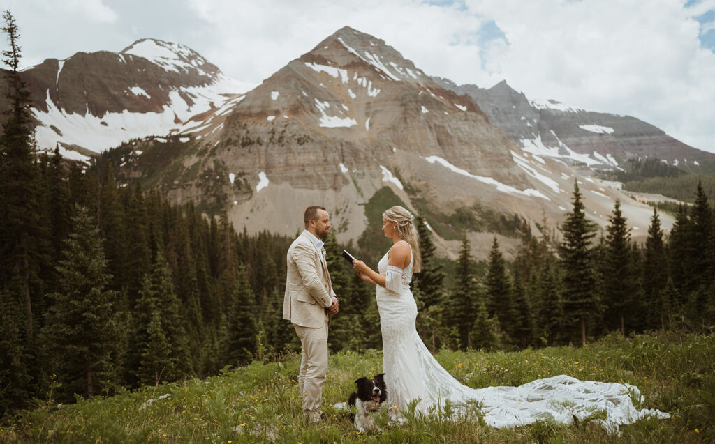 The couple is standing at the base of a mountain with the bride holding her vow book. The groom is wearing a tan suit. Their border collie dog is sitting in the middle of them, smiling. There is green grass and tall mountains surrounding them.