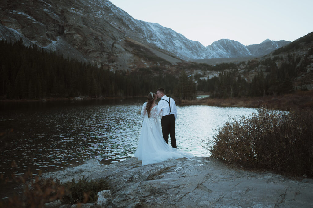 couple is standing on the edge of a lake kissing with their backs to the camera. The sun has already set and there are mountains covered in snow and a lake in the background. The groom has a white shirt and black suspenders and black pants on. The bride has florals in her braided hair.