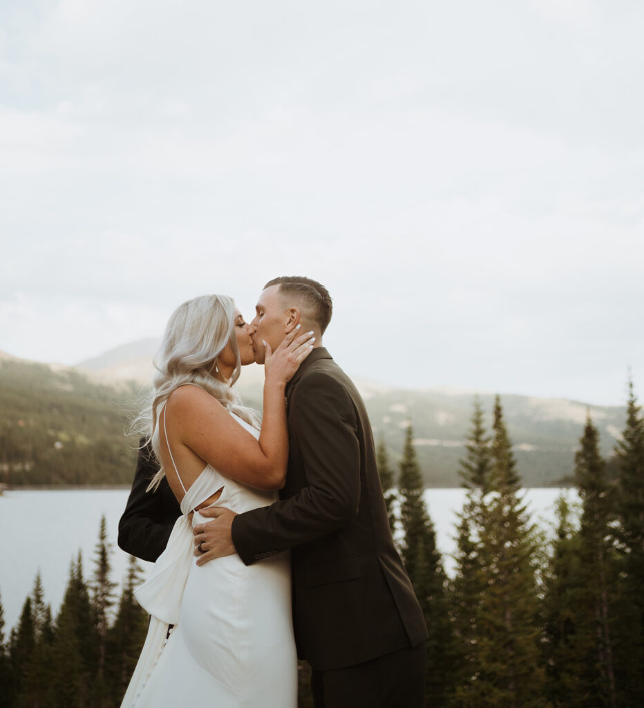 bride and groom are kissing. The bride has her hand on his neck and the groom has his hands around her waist. The bride is in a white wedding dress and the groom is in a black suit. There is a lake in the background and there are pine trees and mountains.