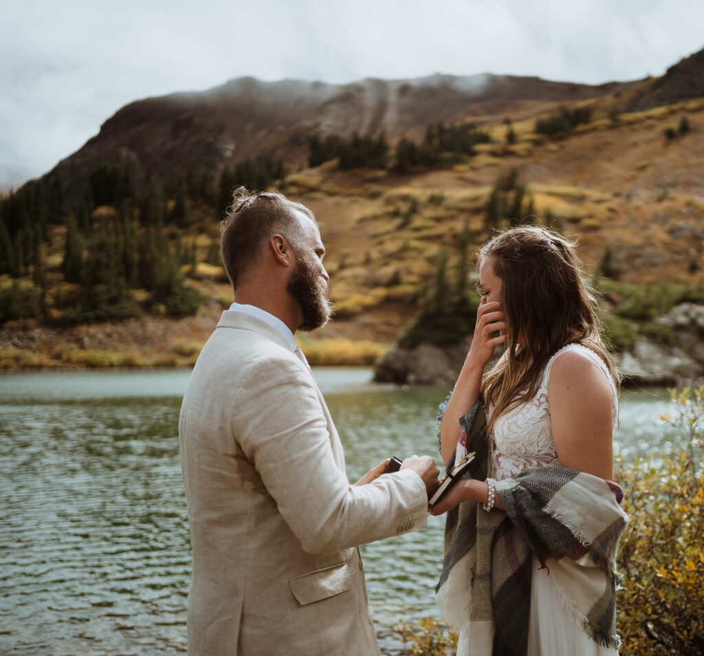 the bride and groom are reading their vows. The bride is crying and wiping a tear away. The groom is opening his vow book. The groom is wearing a tan suit and the bride has a checkered shawl around her waist. The lake has an island with trees in the middle of it, and there are yellow trees in the background.