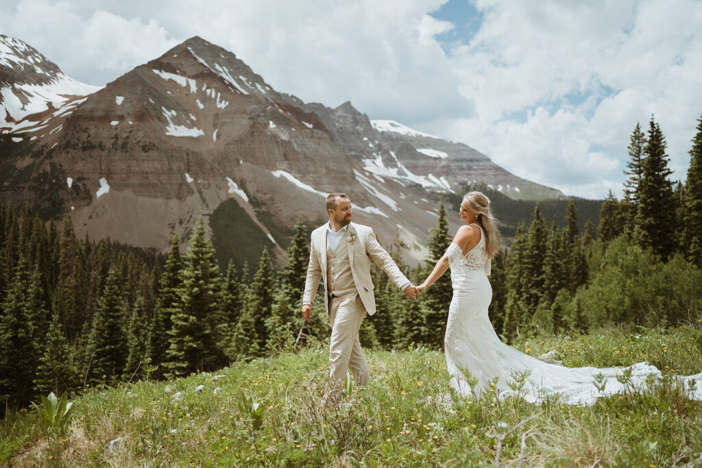 groom is leading the bride through a grassy meadow with wildflowers. There are large mountains in the background and pine trees all around them. The groom is looking back at the bride and the bride is smiling at him.