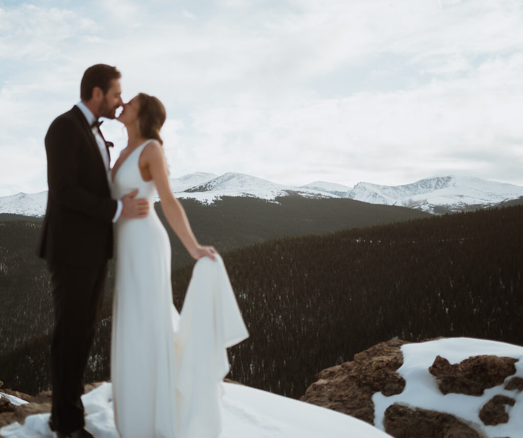 The bride and groom are out of focus and the mountains behind them are in focus. They are standing in snow and the bride is holding the edge of her dress. There are tons of pine trees behind them leading up to the snowy mountains. They are smiling into each other's mouths.