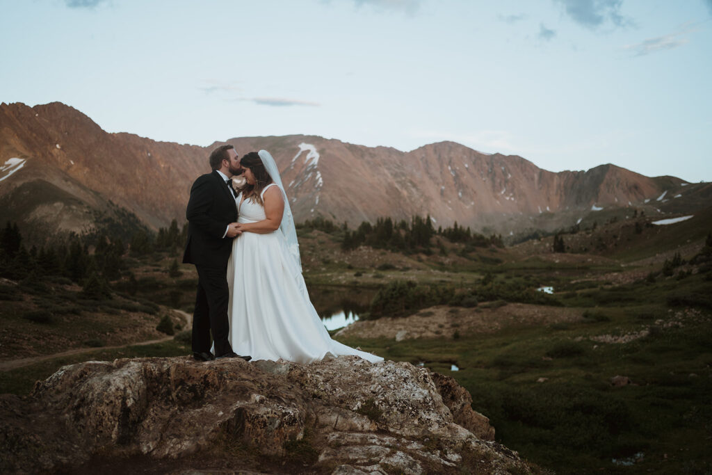 groom is kissing the bride's forehead and they are standing on a large rock overlooking the mountain range. The mountains are stained purple and the couple is holding hands. The grass is green and the pine trees are sparce behind them. They planned an epic colorado elopement on a mountain pass.