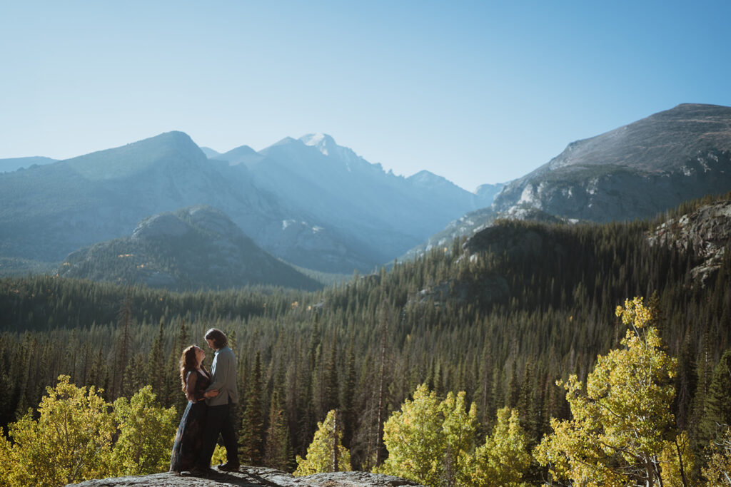 The bride and groom are standing together smiling at each other. there are giant mountains in the background and green trees. There are aspens turning yellow and the couple is standing on a cliff ledge overlooking this.