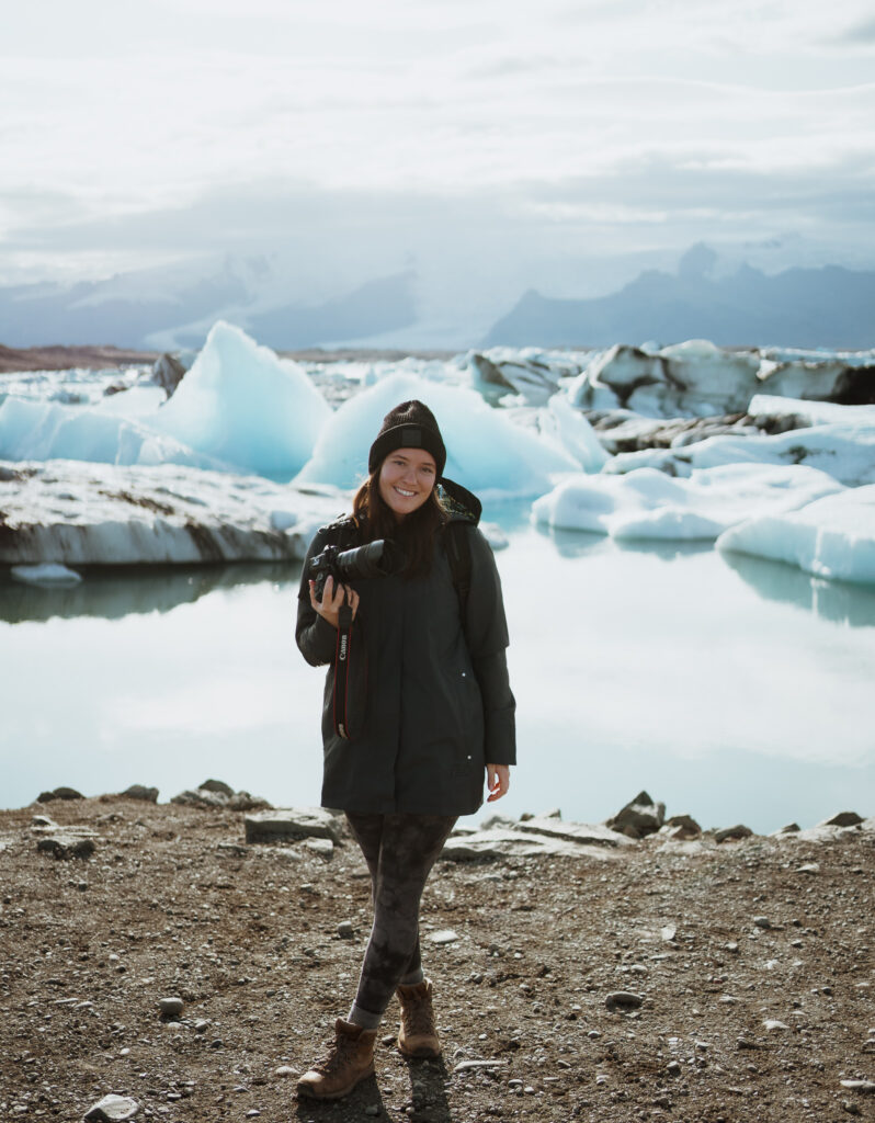 Girl is smiling at the camera. She is holding a camera with ice bergs and a glacial lake in the background. She is wearing a long navy rain coat and grey leggings. She has hiking boots on her feet and a black hat on. She has brown hair and hazel eyes.