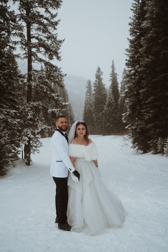 The bride and groom are both staring at the camera without smiling. There is snow all around them and tall pine trees with snow on them. The groom is in a white suit jacket and black pants. The bride is in a white dress with a fluffy coat.