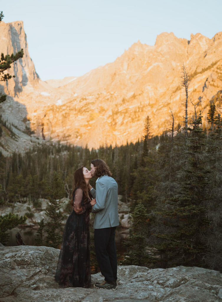 Bride and groom are kissing at sunrise. They are standing on a rock with a mountain that is bright orange. there is a lake behind them. The bride is wearing a non-traditional black dress and the groom is wearing a grey suit jacket and black pants.