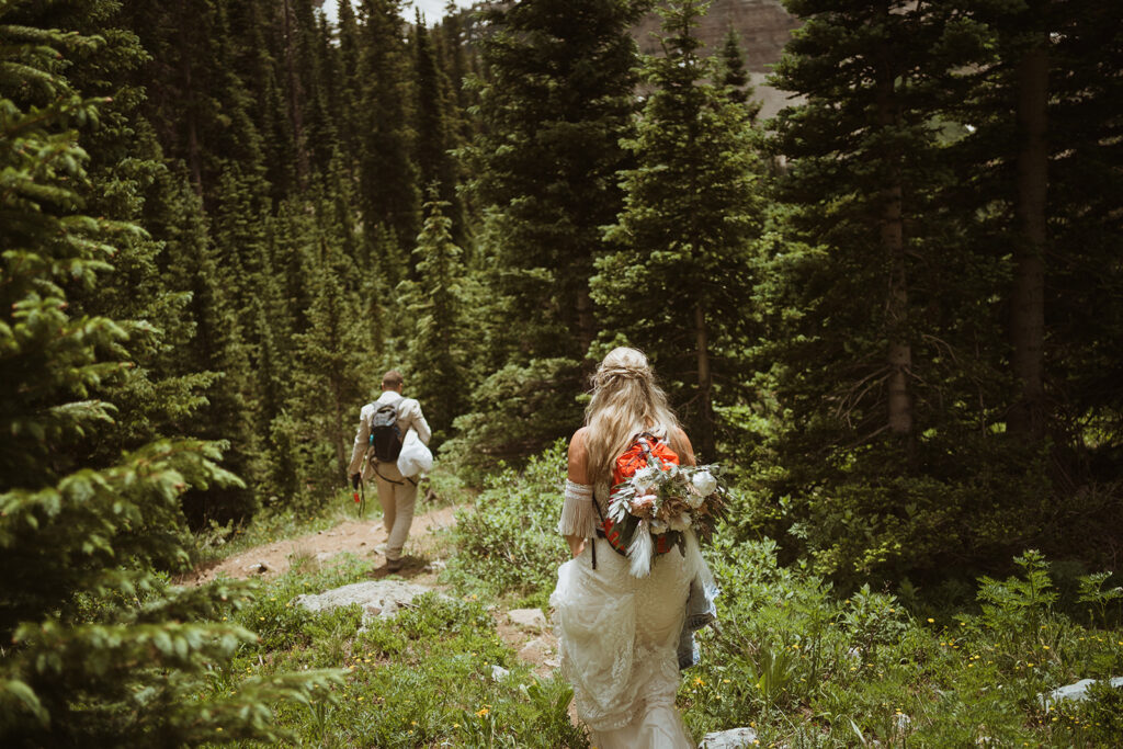 Couple is hiking down a trail. there is greenery and pine trees surrounding them. the groom is walking in front of the bride down the trail with a tan suit on as well as his backpack. The bride is walking up the trail from the groom. She is wearing her wedding dress and has her bouquet attached to her hiking backpack.