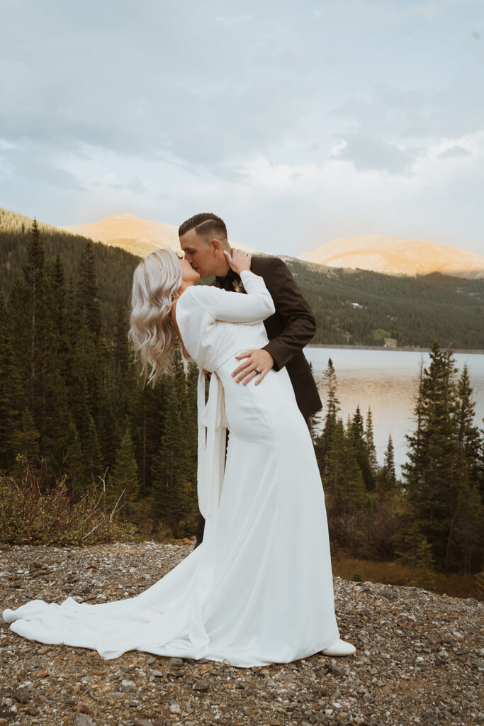 The groom is dipping the bride slightly while the sun sets. There are mountains and pine trees behind them and they are having a colorado elopement at sunset. They are standing on a gravel platform.