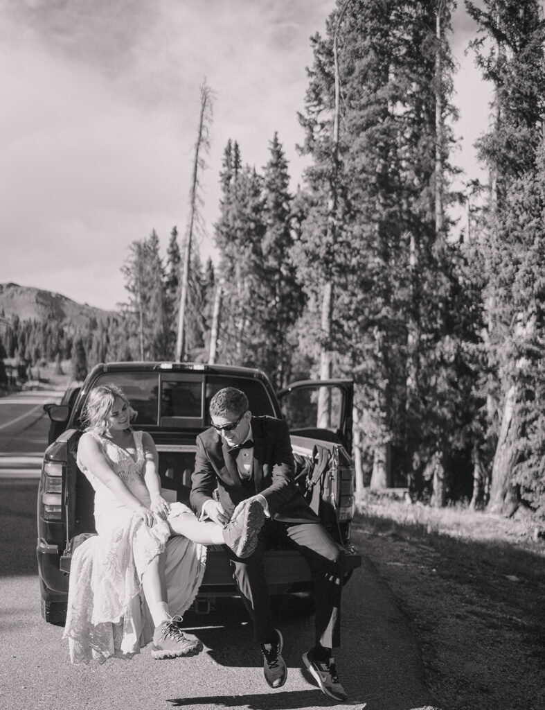 bride and groom are sitting in the bed of a pick up truck. The groom is untying the bride's hiking boot. they are smiling and laughing. There are tall trees and a windy road behind them. The photo is in black and white.
