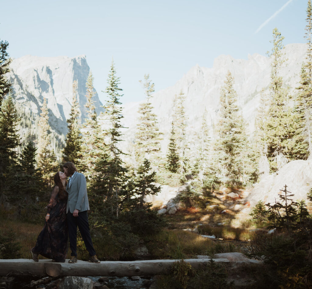 The bride and groom are standing on a log over a river. They are kissing and there are mountains and pine trees in the background. The groom has a grey suit and the bride has on a black dress with flowers. learn how to elope in colorado's national parks.