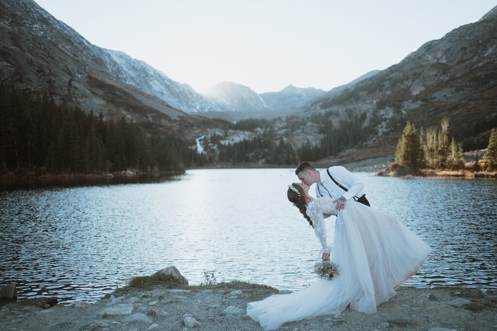 The groom is dipping the bride with a lake and mountains in the background. The groom is wearing a white button up and suspenders. The bride has a multi-colored bouquet. 