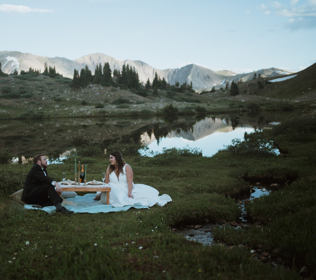 The bride and groom are sitting in green grass. They are sitting on a blue blanket with a table that has champagne glasses and candles. There is a lake behind them with a reflection of the mountains on it. Learn how to elope in colorado with epic activities and picnic locations.
