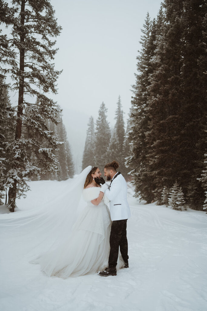 The groom is holding the bride's face and they are both laughing. There are tall pines and snow in the background. The bride has a veil that's blowing in the wind and they are wearing white and black gloves.