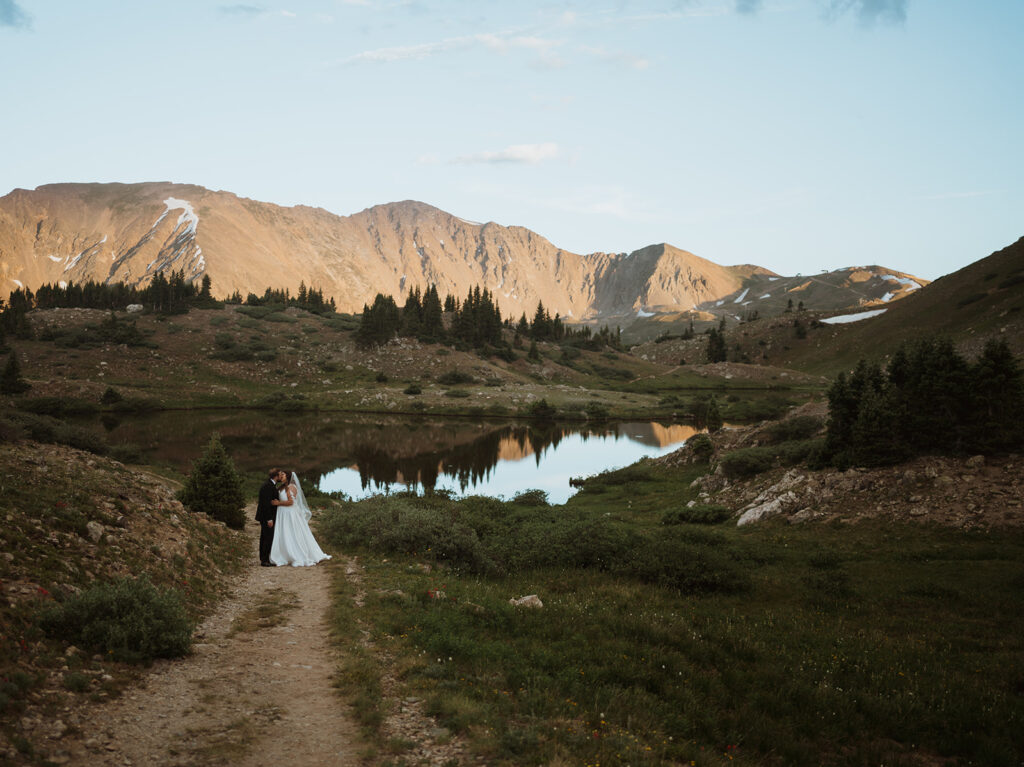 The bride and groom are standing far away and kissing. There are huge mountains that are turning burnt orange in the background with a lake behind them. There is green grass and a trail leading down to them. 