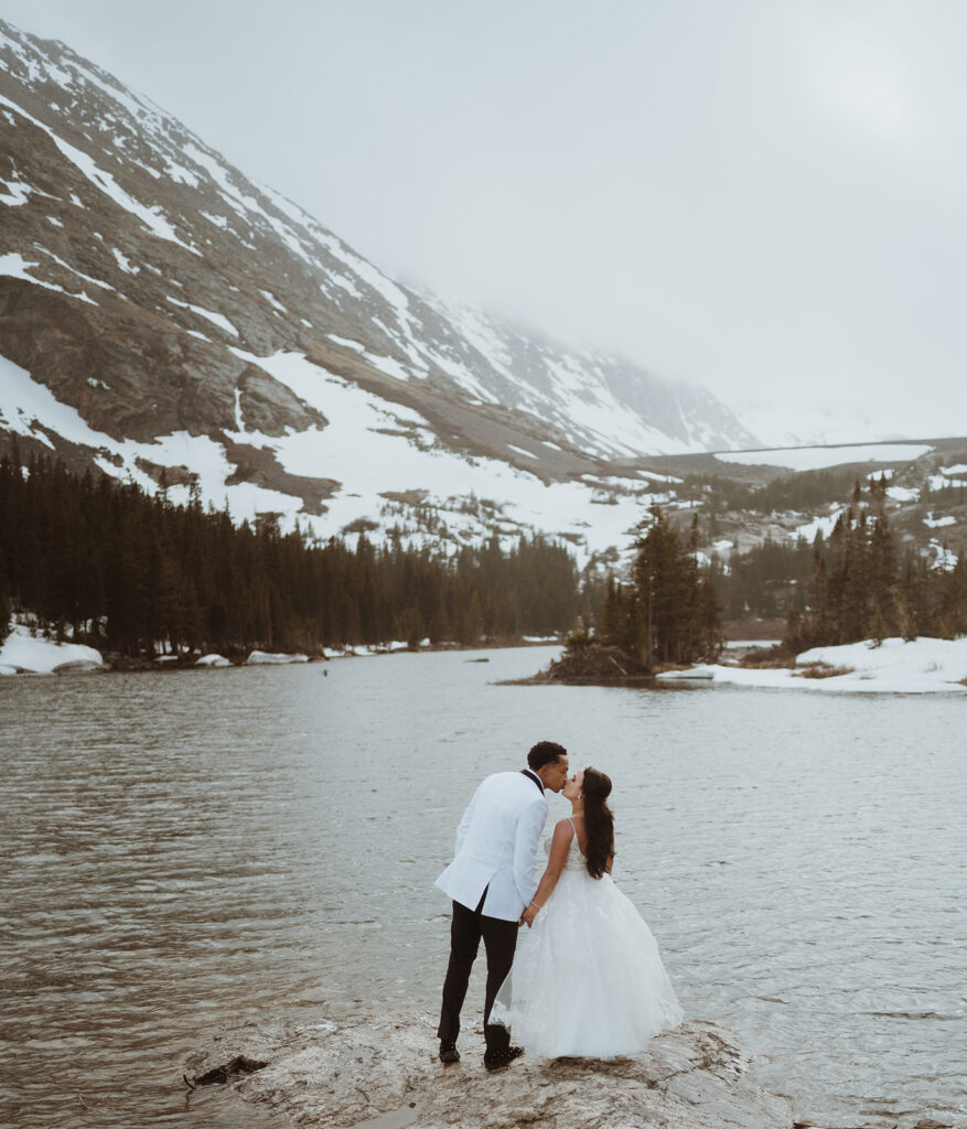 Couple is holding hands and standing at the base of a lake and kissing. There are mountains covered in snow and fog in the background. The groom is in a white suit jacket and black pants. The bride is in a white wedding dress.